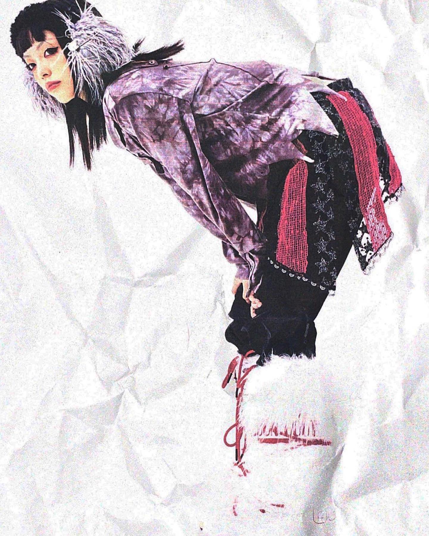 the image showcases a female model in a striking ensemble characteristic of japanese alternative fashion styles such as visual kei or harajuku. she is wearing a tiedye purple shirt with distressed edges, layered over black pants. the outfit includes a noticeable red and black accessory piece draped around her waist and  fluffy white boots with red laces. her hair is long and black with a feathered headband. the background is a textured, crumpled white paper which complements the overall edgy and artistic feel of the photograph. the overall aesthetic suggests a blend of punk, grunge, and other alternative fashion elements. the model's pose and styling contribute to a strong sense of attitude and individual expression.