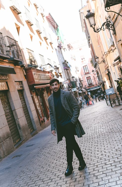 a man in a coat and turtleneck stands on a cobblestone street next to buildings. he is illuminated by light with shadows cast around him.