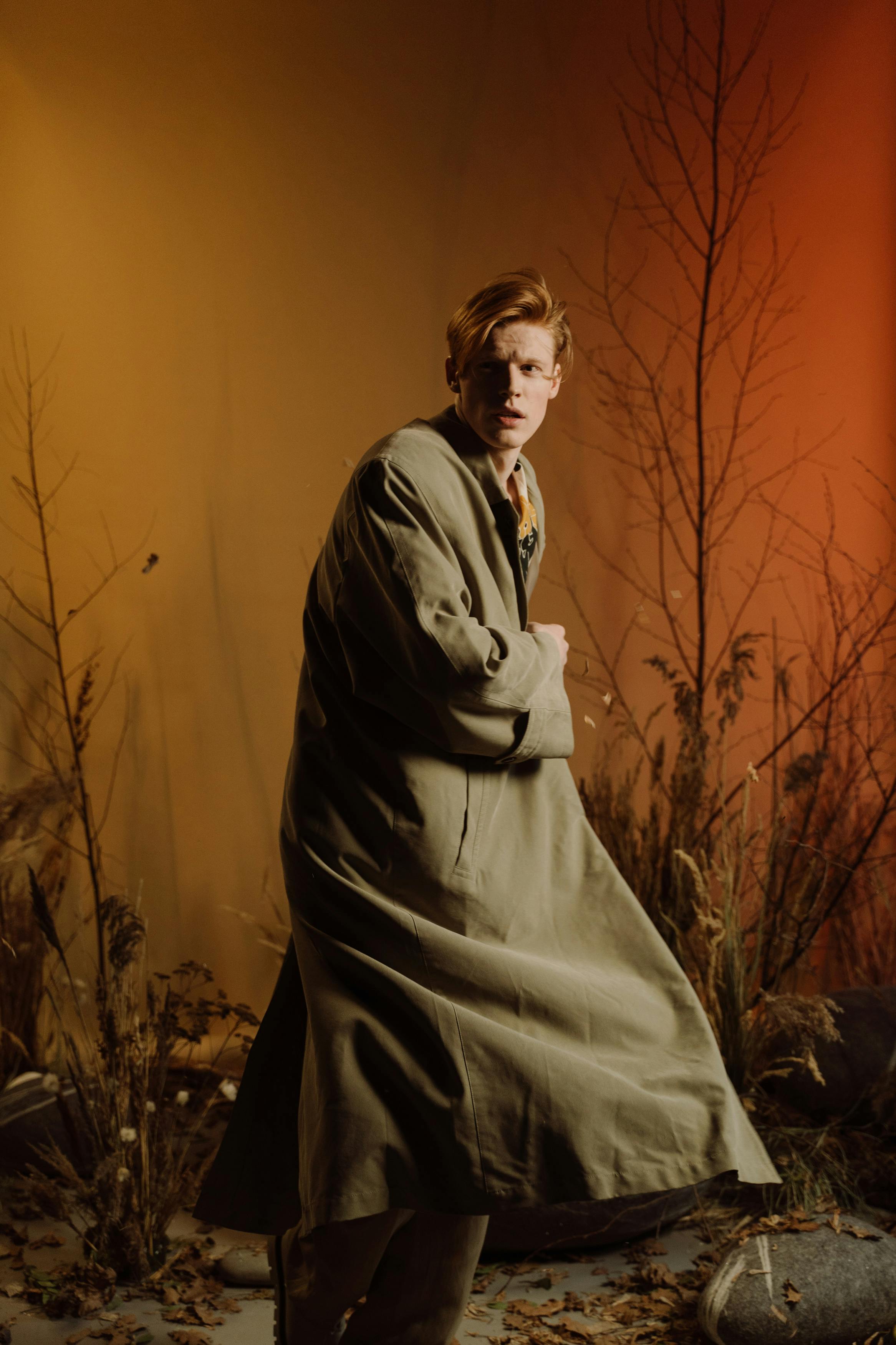 a young, paleskinned redhead man with freckles is standing in a studio shot against a backdrop of dry, autumnal plants and branches. he is wearing a long, loosefitting khaki or beige trench coat, and his expression is serious and somewhat intense. the lighting is dramatic and moody, with warm, amber tones dominating the scene, creating an artistic and stylistic feel. the overall impression is one of fashion photography, highlighting the model's clothing and the evocative atmosphere.