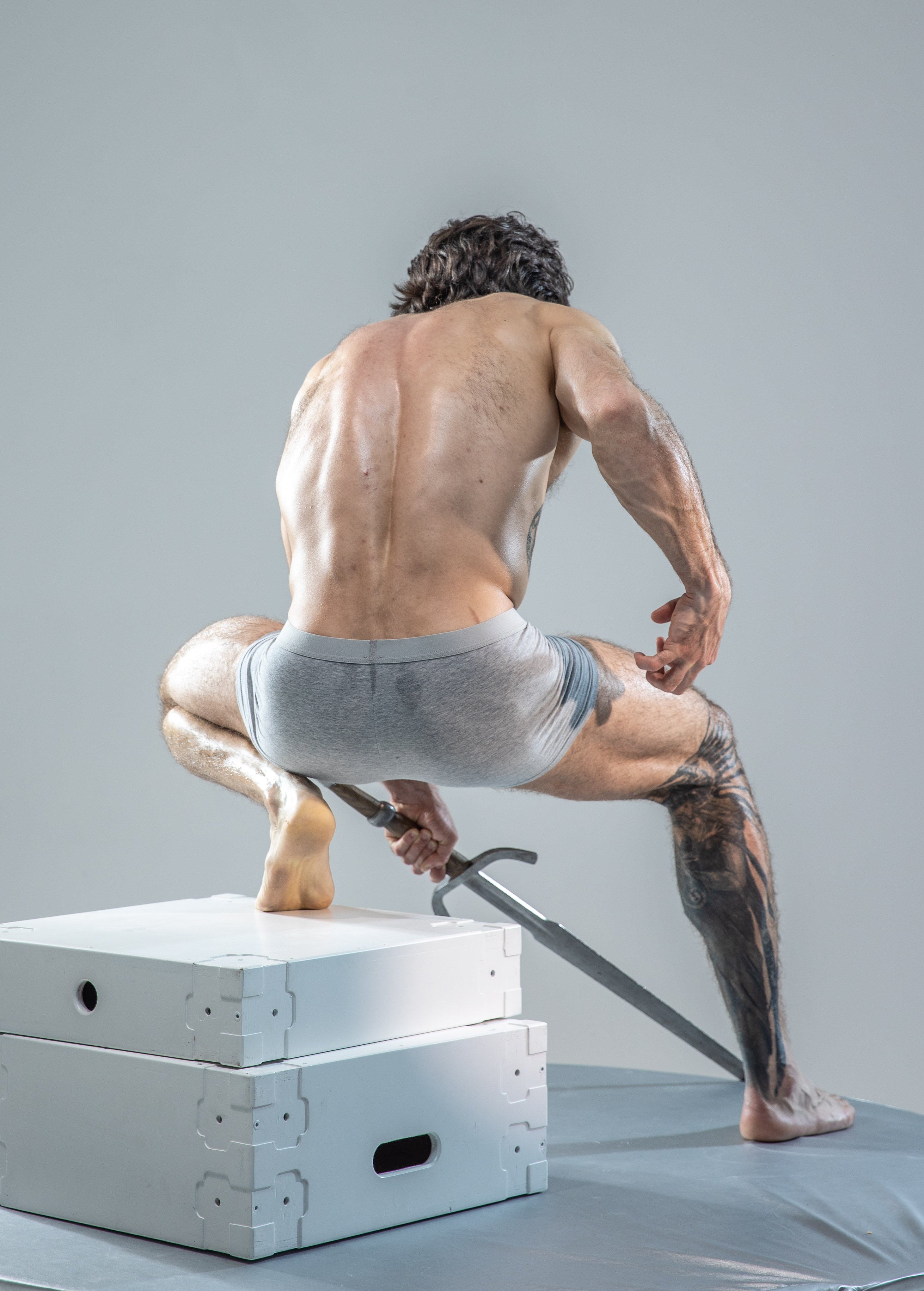 a muscular man with tattoos is posed dynamically on two white boxes against a plain gray background. he is shirtless, wearing only gray underwear, and holds a sword in his right hand, the blade extending in front of him.  the lighting is soft and even, highlighting the man's muscular physique. the overall image gives a sense of action and power.