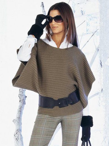 a woman in winter fashion, wearing sunglasses, a brown poncho cinched with a wide black belt, plaid pants, and gloves, is standing against a wintry background. she has long dark hair and is wearing hoop earrings. she is wearing a white blouse under the poncho. her left hand is holding the sunglasses while her right hand is holding a purse with a furry cuff.