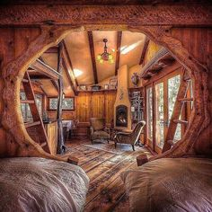 this image shows a rustic and cozy interior, possibly a treehouse or cabin, with a wooden design. the space features two beds in the foreground, a small dining area with a table and chairs, a fireplace, and comfortable armchairs. a ladder is used to access a higher level. the walls, ceiling, and floor are made of wood, creating a warm and inviting atmosphere. the room is illuminated by a chandelier and natural light through a window. the overall impression is one of comfort and charm.