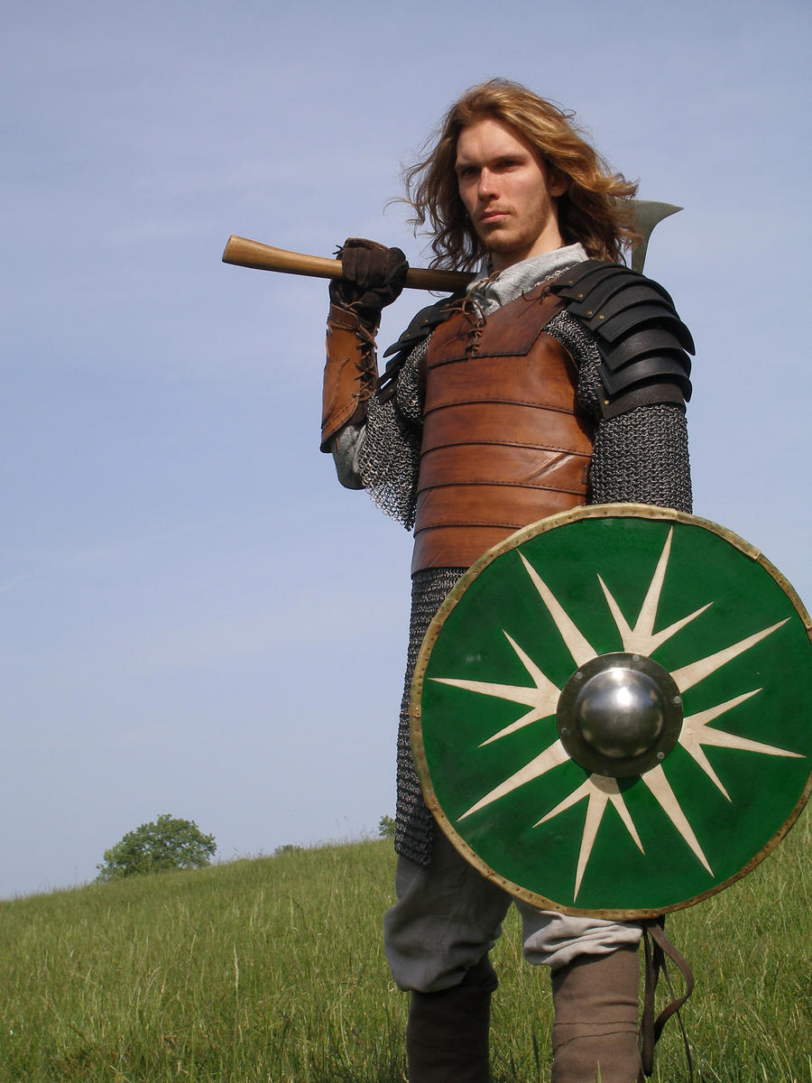a young man dressed in medieval warrior garb stands in a grassy field against a clear blue sky.  he wears a leather jerkin over chainmail, leather arm guards, and holds a large axe over his shoulder. he also carries a round green and white shield with a central boss. his long, light brown hair is somewhat loose. the overall impression is one of historical reenactment or fantasy roleplaying.