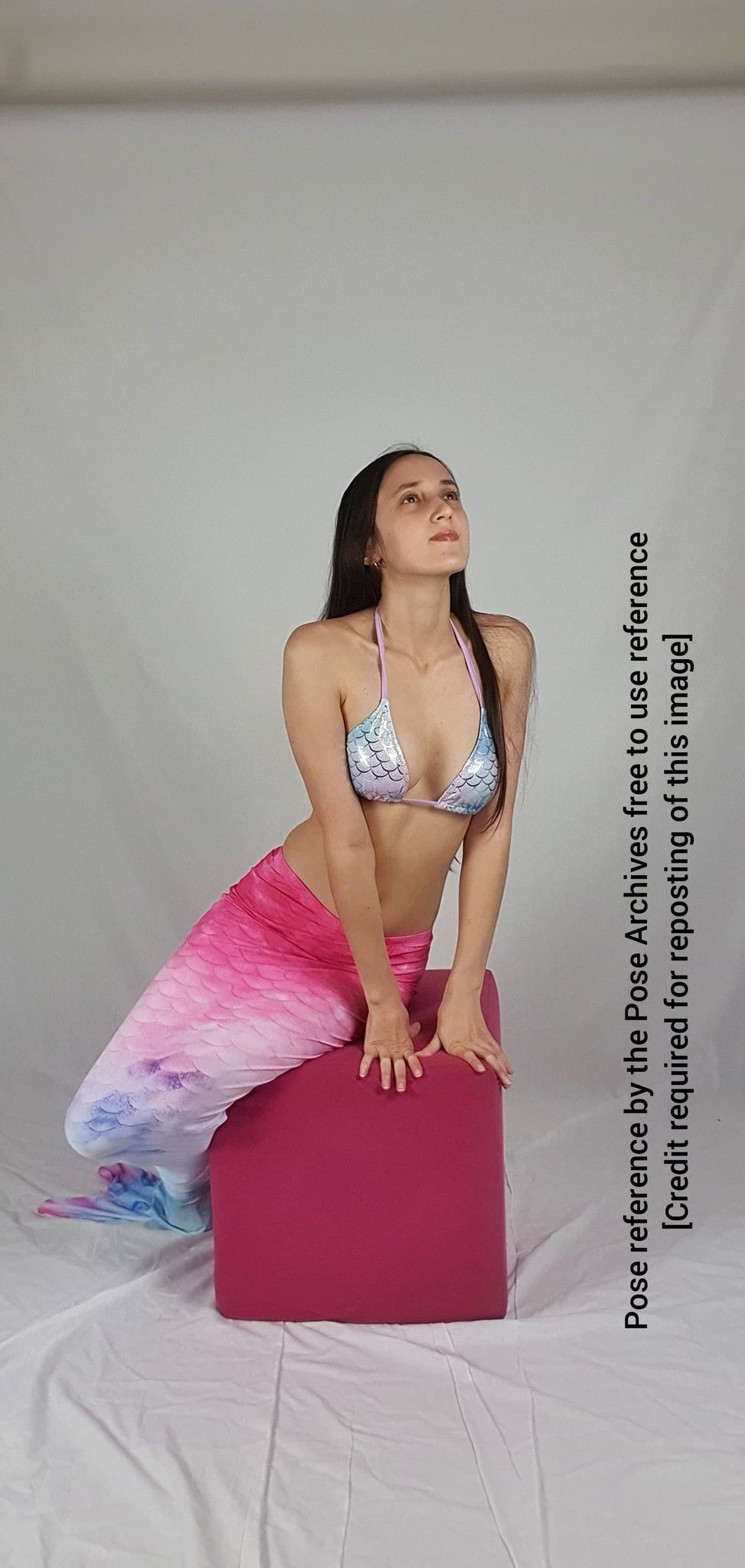 a woman with long black hair is posing in a mermaid costume. she is wearing a bikini top with a mermaid scale pattern and a pink and purple ombre mermaid tail. she is sitting on a pink cube and looking up. the background is a white wall. the lighting is soft and the overall tone of the image is light and airy.