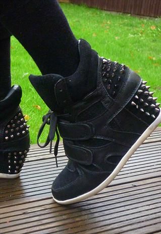 closeup shot of a person wearing black, spiked sneakers. the shoes have a platform sole, two velcro straps and black shoelaces. the shoes also have a row of spikes around the ankle area. the person is wearing black socks. the background is of grass and a wooden surface.