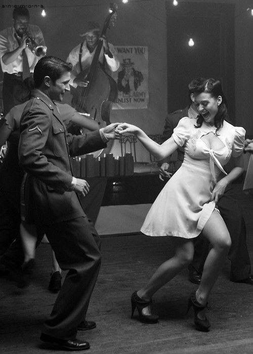 a man and woman are dancing in a room. the woman is wearing a white dress and the man is wearing a suit jacket and slacks. the woman is smiling and the man has a serious expression. the woman is holding her hand up as if she is about to spin. the man is holding her hand with one hand and