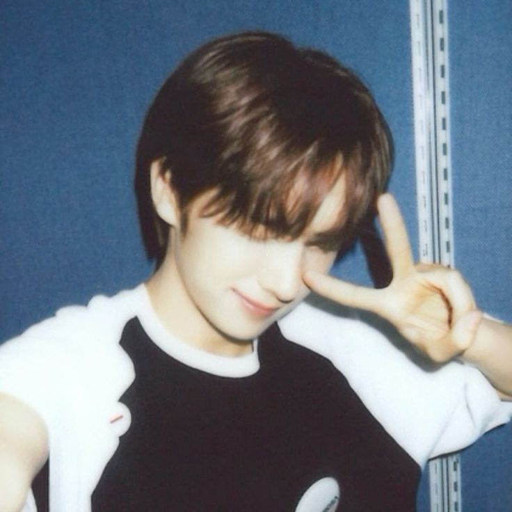 a young asian boy with brown hair and bangs is making a peace sign with his hand in front of his eye. he is wearing a black and white tshirt and smiling. he is looking at the camera. the background is blue and striped. the image is taken from a closeup angle. the image has a grainy, vintage feel. the image is probably a photo taken with a polaroid camera.