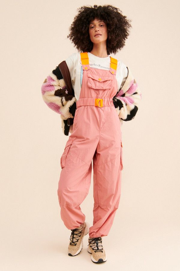 a fashion model with curly afro hair is wearing a pair of dusty rose colored cargo overalls with orange straps and a beige tshirt underneath. she is also wearing a patterned brown and cream colored fuzzy jacket and beige chunky sneakers. the model is standing against a light beige background.