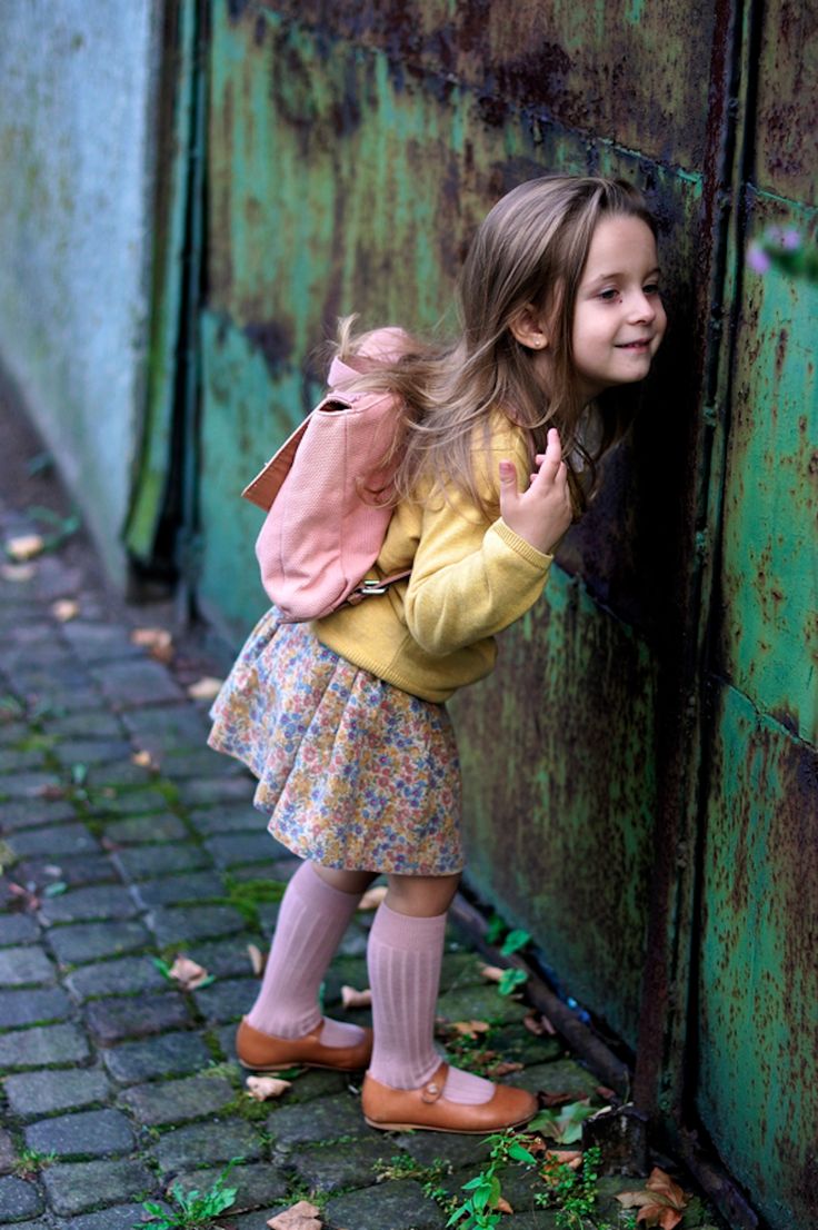 a young girl with a pink backpack and floral skirt is looking through a rusty gate. she is wearing a yellow sweater, pink socks, and brown mary jane shoes. she is standing on a brick path.