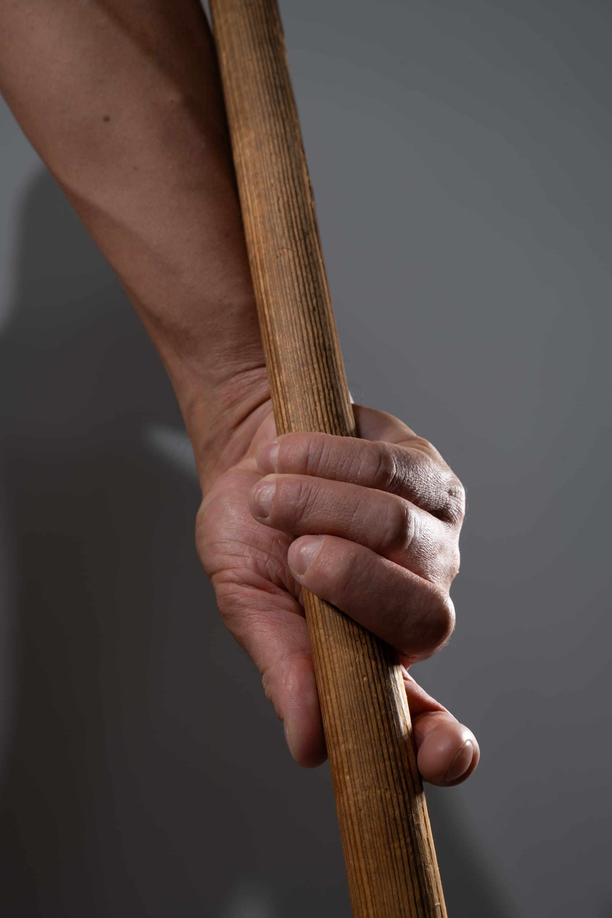 a closeup of a man's hand gripping a wooden stick against a gray background. the hand is strong and weathered, suggesting age and experience.  the stick is smooth and cylindrical, its surface showing signs of wear. the lighting highlights the texture of the hand and wood, creating a sense of realism.