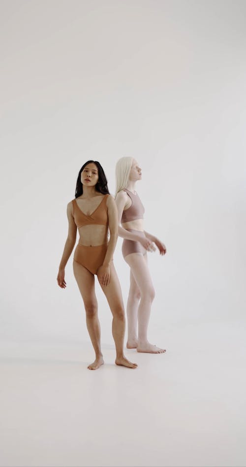 two women, one with brown skin and one with albinism, stand sidebyside in minimal peachtoned underwear against a plain white background. they are positioned slightly turned to the side, giving a full view of their bodies. the women appear confident and at ease, conveying a message of body positivity and inclusivity.  the lighting is soft and even, highlighting their bodies and the subtle differences in their skin tones. the overall aesthetic is clean, minimalist, and focused on celebrating diverse beauty.