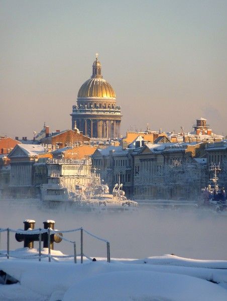 a stunning winter scene in saint petersburg, russia. the iconic golden dome of saint isaac's cathedral is prominently featured in the background, rising above a line of snowcovered buildings along the neva river. a light fog hangs over the water, adding to the serene and magical atmosphere.  the foreground shows a snowcovered embankment with a metal railing and bollards. boats are visible in the misty river. the overall impression is one of quiet beauty and cold, crisp winter air.