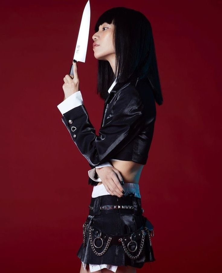 a woman with short black hair and bangs is holding a kitchen knife in front of her face. she is wearing a black jacket, a black skirt with a chain belt, and a white top underneath. she is looking at the knife with a serious expression on her face. the background is a solid red.