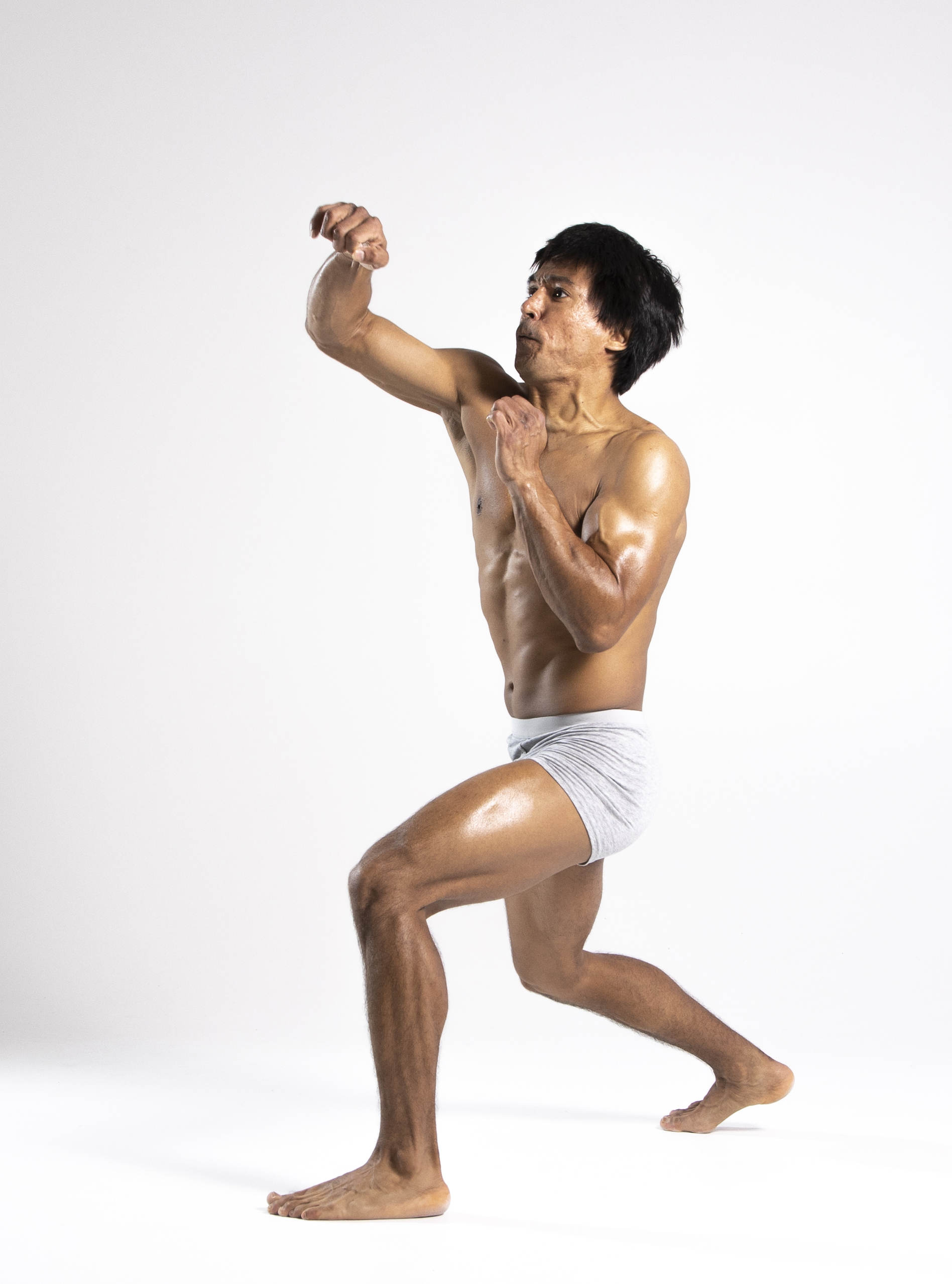 a shirtless man in white underwear is in a boxing stance on a white background. he is barefoot, muscular and punching with his left arm extended.