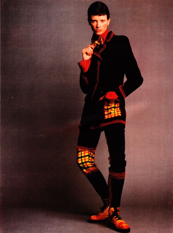 a full shot features a model standing with an androgynous appearance and short hair, dressed in a distinctive fashion ensemble against a mottled, purplishgray backdrop. the model is wearing a black suit with red trim, which includes a blazer and matching trousers. the suit is notable for its embroidered details, including plaid patches on the knees of the trousers and on the blazer's pockets. the model's attire includes a red shirt under the blazer. the look is completed with a pair of leather shoes with red accents. the model's pose is relaxed, with one hand touching their collar and the other resting in their pocket. the overall aesthetic combines classic suit tailoring with unique, embellished elements.