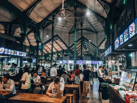 the image shows the bustling interior of lau pa sat, a historic food court in singapore.  the victorianera cast iron structure of the building is prominent, with its intricate metalwork forming a high, open ceiling. many people are seated at tables, enjoying food from various stalls. the atmosphere is vibrant and lively, showing a diverse crowd of people enjoying a meal. various food stalls are visible along the sides of the building, showcasing the diverse culinary offerings. the overall scene captures the energy and culture of a typical night at this popular singaporean food court.