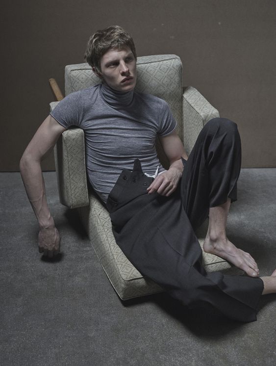 a young man in a grey shirt and black pants sits on a couch in a studio. he is casually leaning back and is looking at the camera. the room is simple and minimalistic.