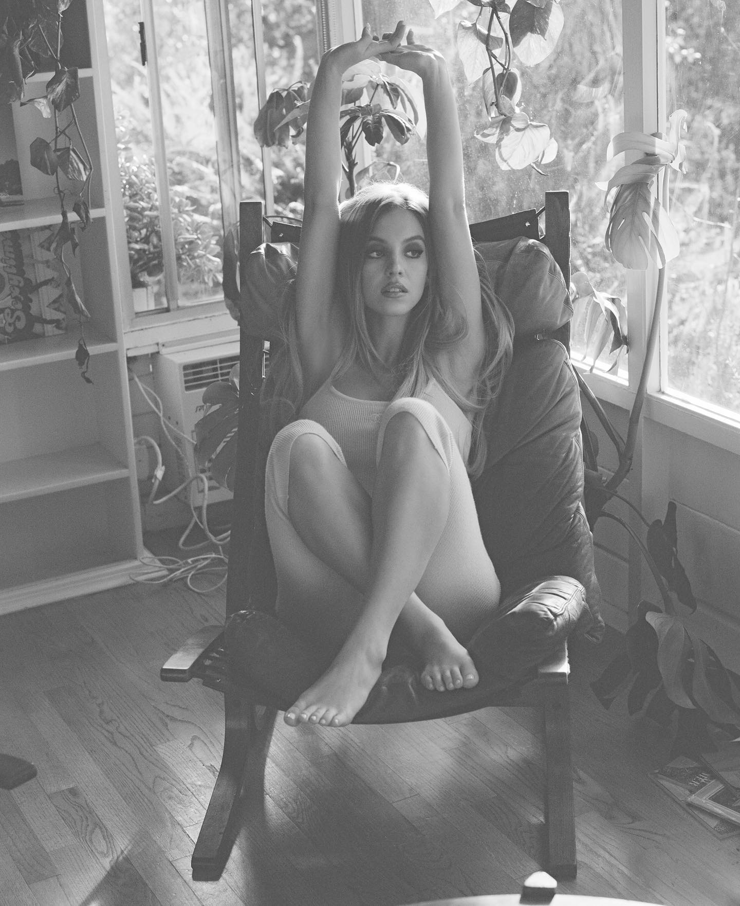 a black and white, fullshot photograph depicts a young woman sitting in a leather armchair. she is barefoot and has her legs crossed at the ankles. her arms are raised above her head, suggesting relaxation or stretching. she is wearing a simple, lightcolored top and what appears to be lightcolored shorts or a skirt. the setting is indoors, in a room with large windows that let in natural light. greenery and plants are visible both inside and outside the window.  the overall mood of the image is calm, peaceful, and somewhat sensual. the lighting and composition emphasize the woman's relaxed posture and the natural beauty of the setting.