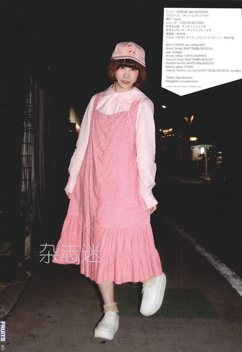 a young woman is posing for a japanese street style magazine. she's wearing a pink floral dress over a white collared shirt.  she's accessorized with white platform sneakers and a white baseball cap.  the photo was taken at night on a city street.