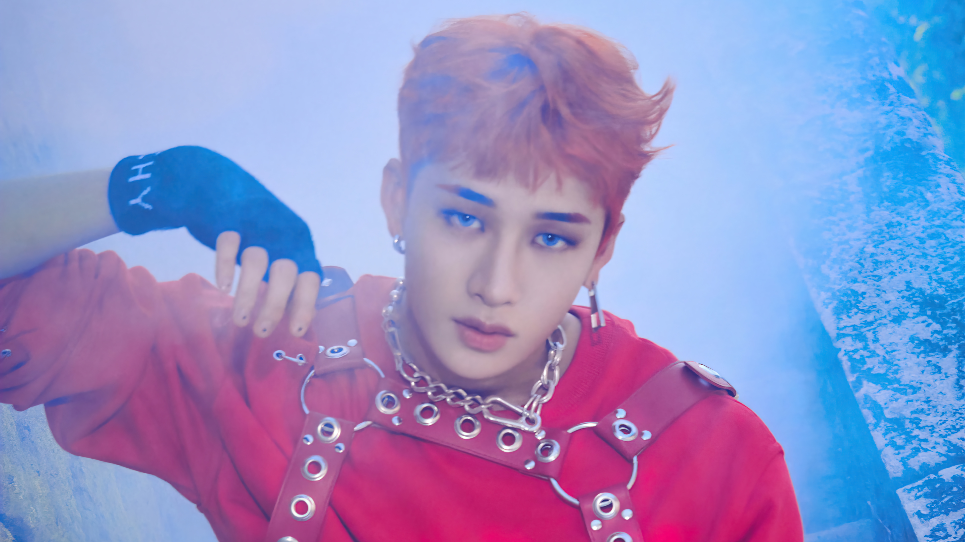 a close up headshot of a young man, with orangered hair, wearing a red shirt with leather straps, a silver chain necklace, and a black fingerless glove. his blue eyes are focused on the camera, and he is surrounded by a soft blue haze.