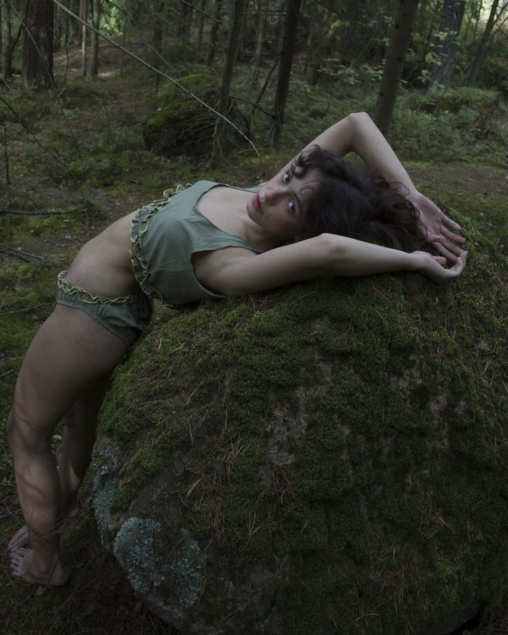 a woman in green swimwear poses in a forest. she is leaning back on a large mosscovered rock. the background consists of trees and forest floor.