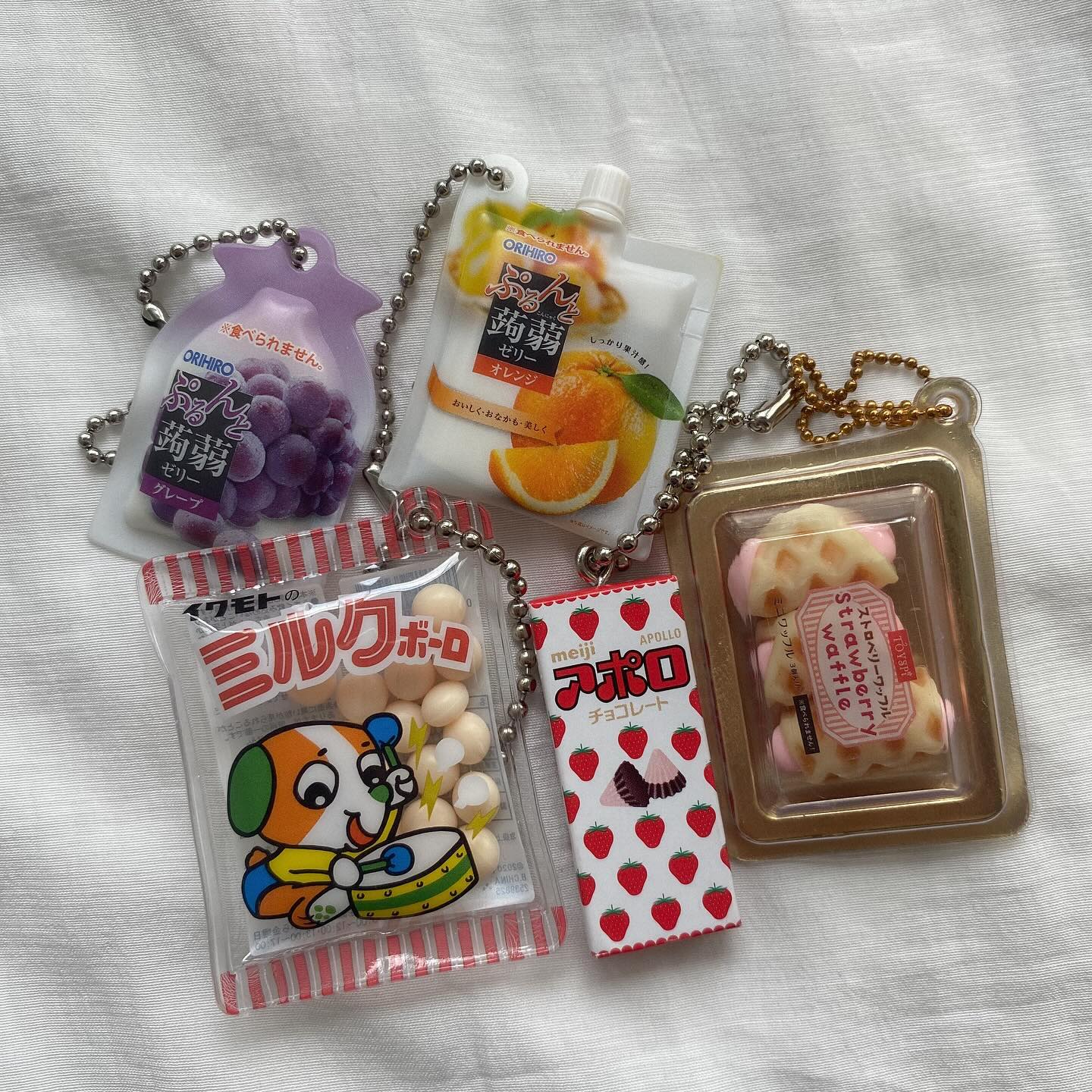 the image shows a variety of japanese snack keychain charms. there are two fruit jelly charms in pouches with nozzles, one grape and one orange flavor. there is also a "milk bolo" candy charm, an apollo chocolate charm, and a strawberry waffle charm.