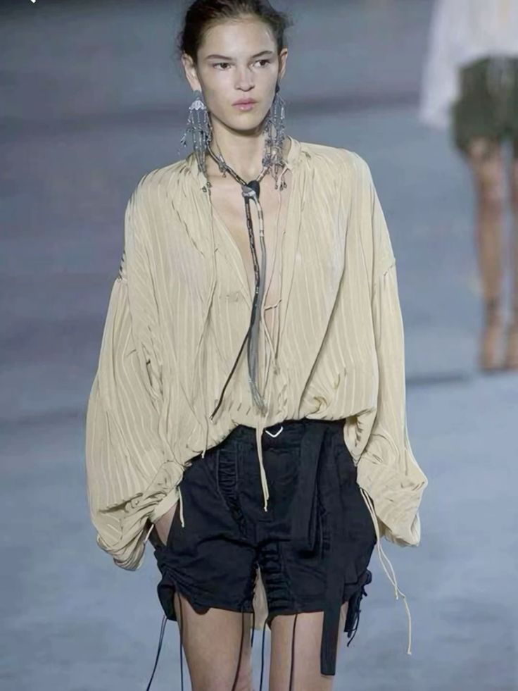 a woman model poses on a runway wearing a cream, loosefitting blouse, black shorts, a long silver necklace, and dangling silver earrings. the background is gray and there are other models in the distance.