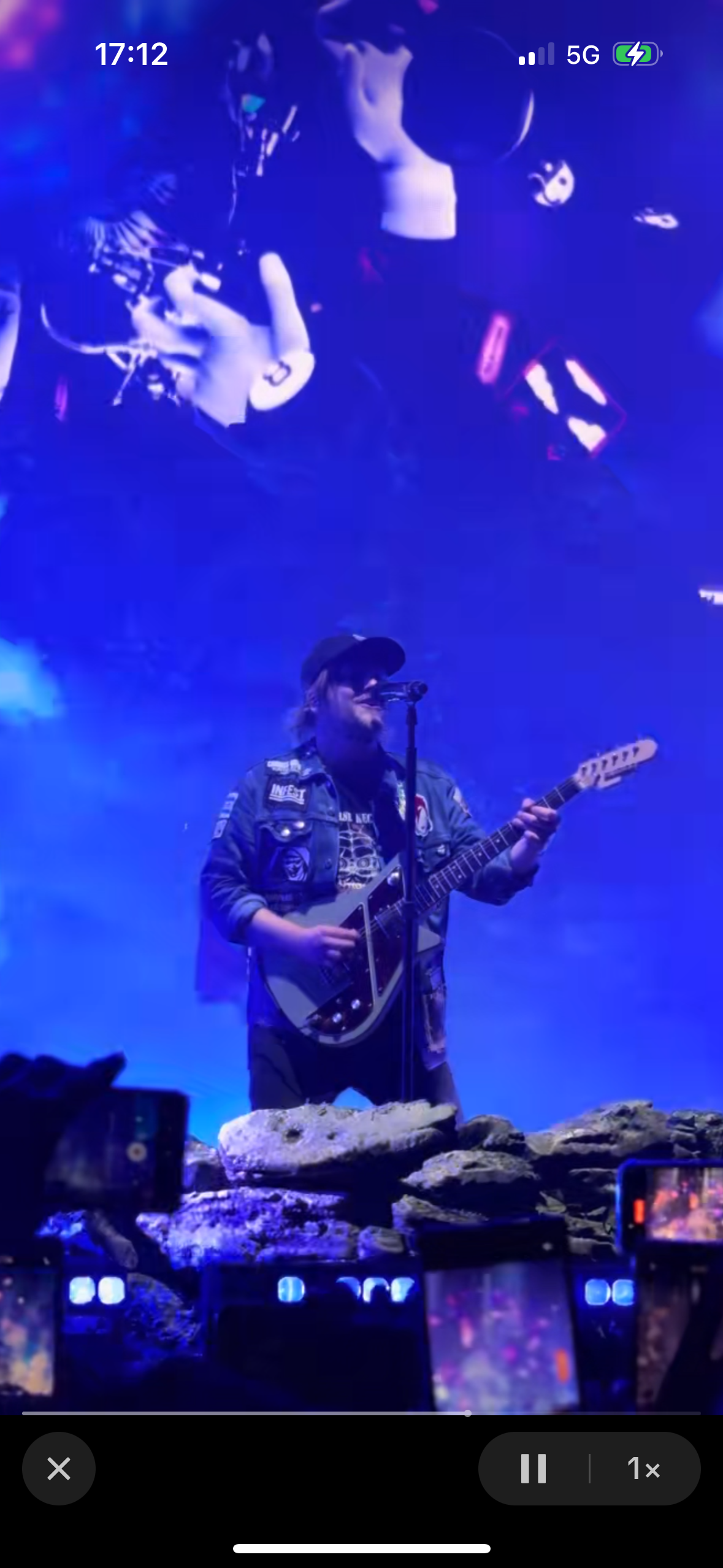 a rock musician is singing and playing a guitar on a stage with blue lighting. the audience is out of frame but the stage is lit up by stage lights. the musician is wearing a denim jacket with patches on it.