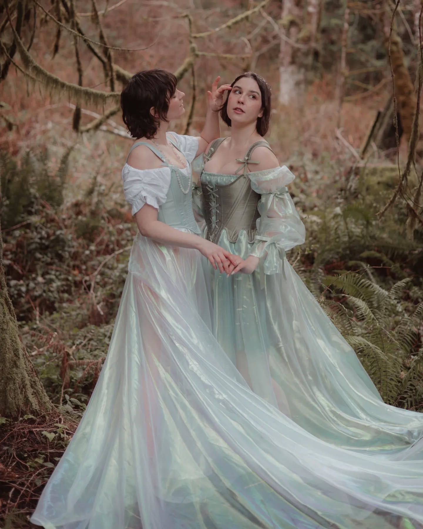 two women in long, flowing, pastel green gowns stand in a forest.  the gowns are sheer and iridescent, giving an ethereal quality. one woman has her hand on the other's face.  the setting is a dark, mossy forest with branches reaching up around them. the overall mood is romantic and fantastical, like a scene from a fairytale.