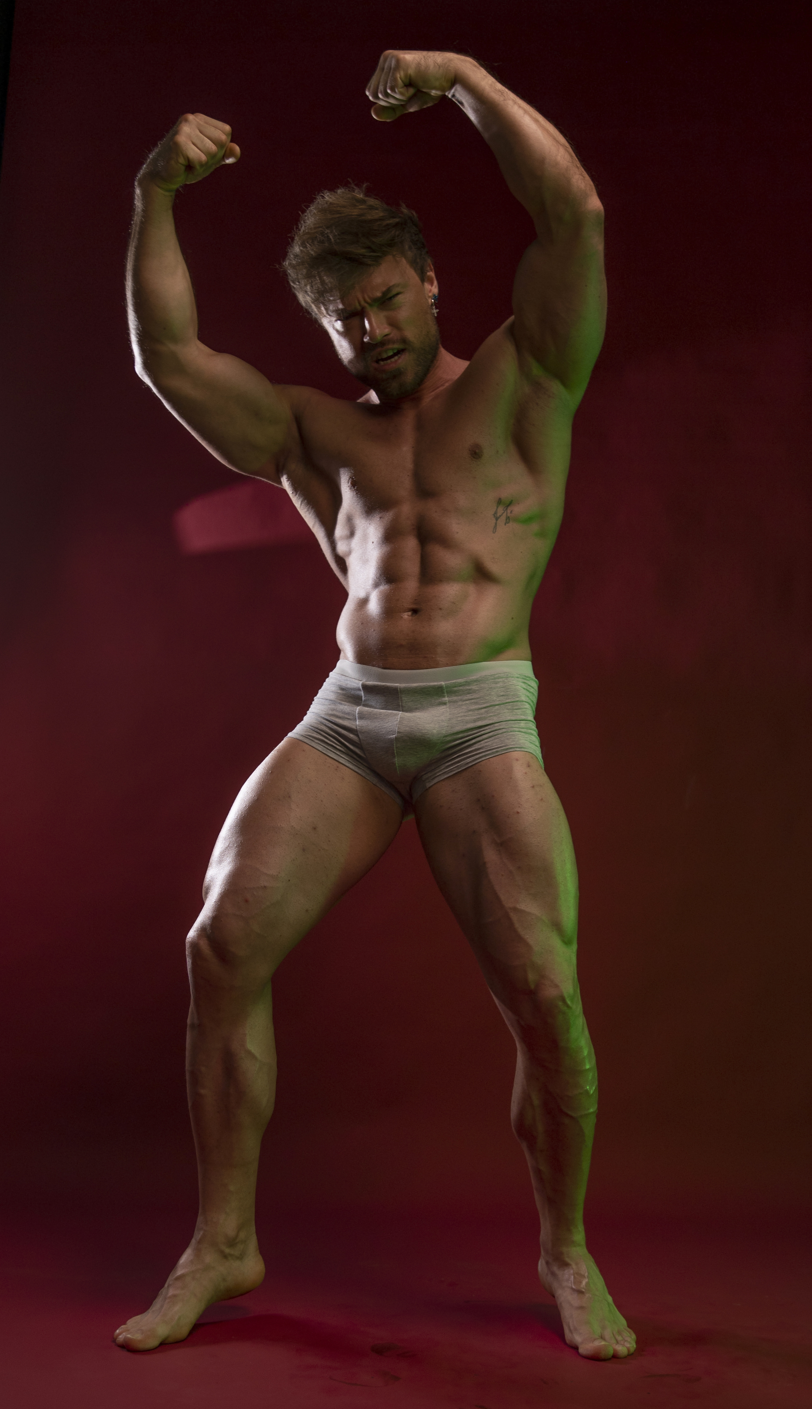 a muscular man is posing in a studio. he is flexing his biceps and showing off his abs. he is wearing underwear and has a tattoo on his torso. the background is red and the lighting is green.