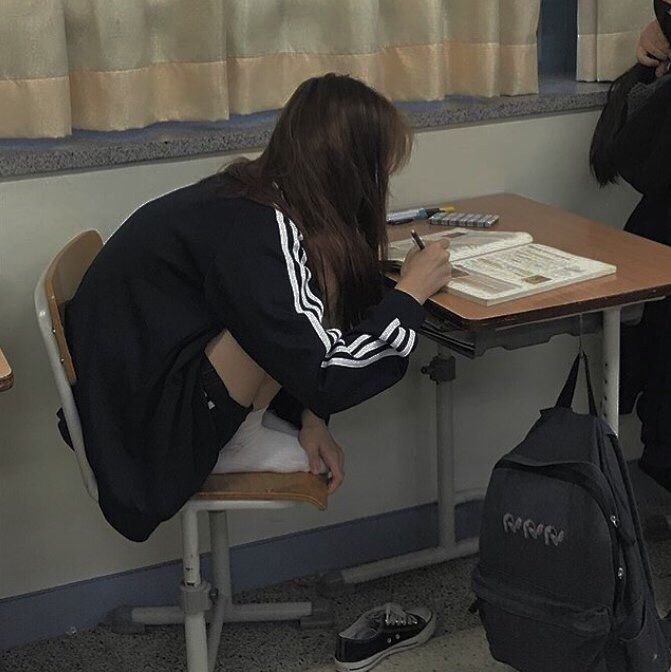 a girl is sitting on a chair in a classroom while writing in a notebook on a desk. she is wearing a black jacket with white stripes on the sleeves and a dark skirt. her hair is long and brown. a black backpack is hanging on the side of the desk, and a shoe is on the floor. curtains are behind the desk.