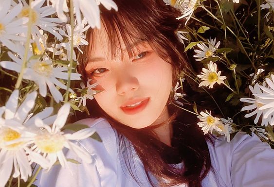 a woman takes a selfie while lying in a field of white and yellow daisies. the sun is shining brightly, illuminating her face and the flowers around her. she is wearing a white shirt and has a happy expression on her face.