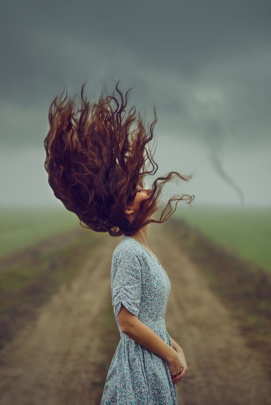 a woman in a blue and white floral dress is standing on a dirt road in a field. her long brown hair is blowing in the wind as she looks up at the stormy sky. the sky is filled with dark, ominous clouds.