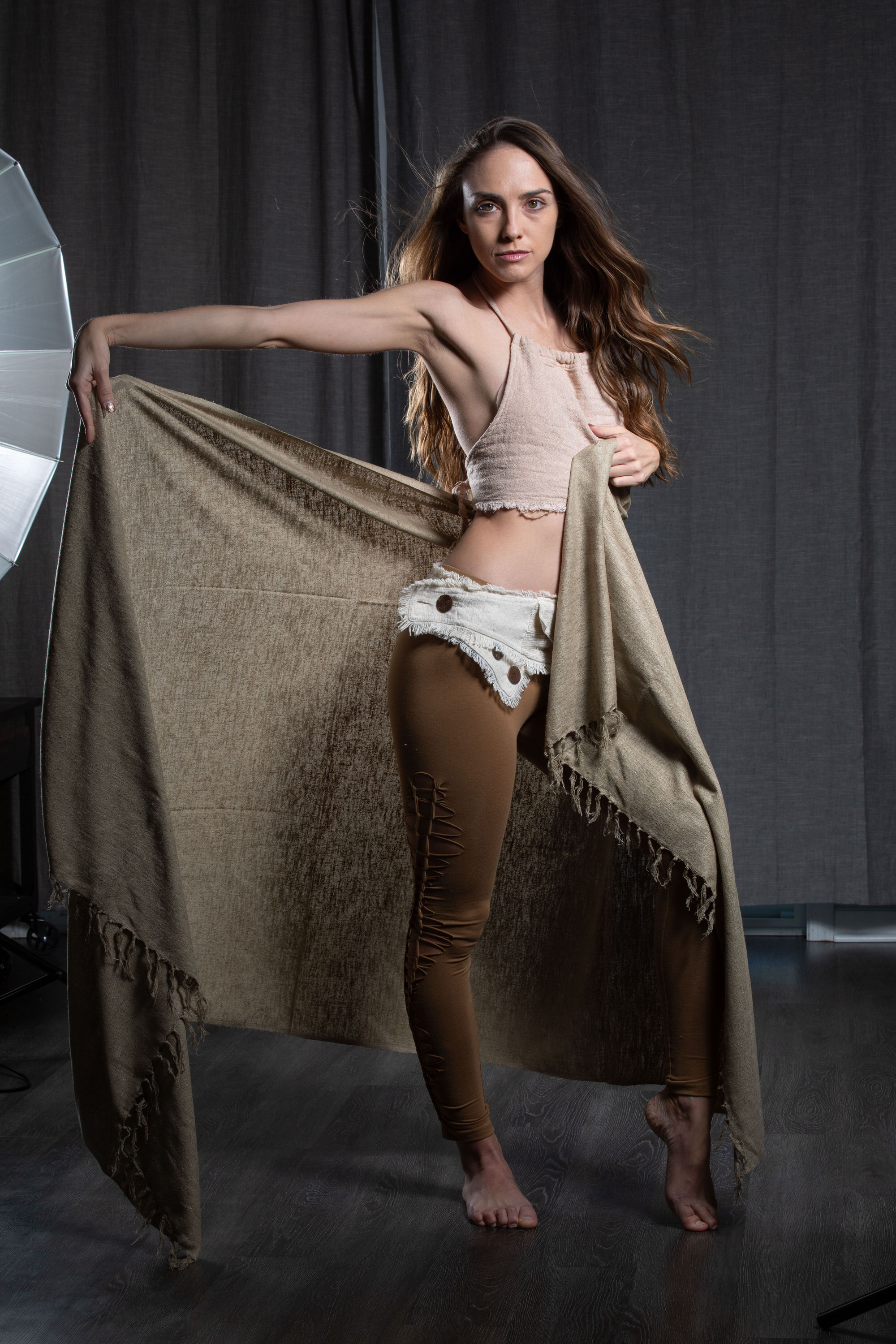a woman with long brown hair stands barefoot in a photography studio. she wears a beige crop top and unique brown shorts. she dramatically holds a large, textured brown cloth, letting it drape across her body and pool on the floor. her pose suggests movement and grace, hinting at a dance background. the lighting is soft and diffused, likely from the softbox partially visible on the left. the overall aesthetic is artistic and expressive.