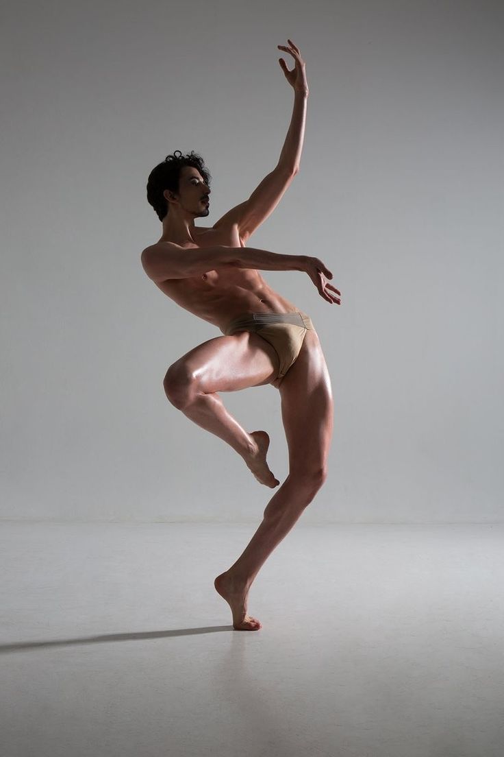 a male ballet dancer poses in a studio, he is wearing a pair of brown underwear and his arms are extended, his right leg is bent in front of him with his toes on the floor. his left leg is extended in the air and his face is facing away from the camera.  he has a very muscular build.