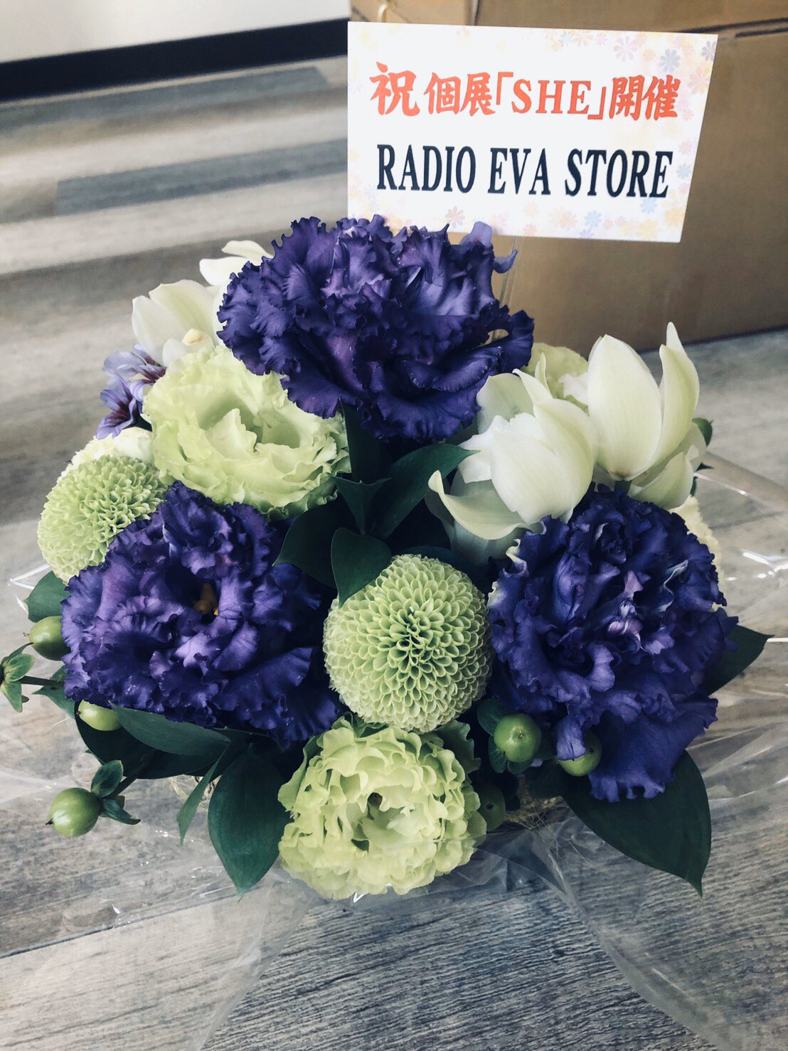a vibrant floral arrangement featuring purple and green flowers is captured in the image. the arrangement, possibly a gift for a store opening, is accompanied by a message in asian characters, suggesting a celebratory occasion. the bright, artificial lighting enhances the vivid colors of the flowers.