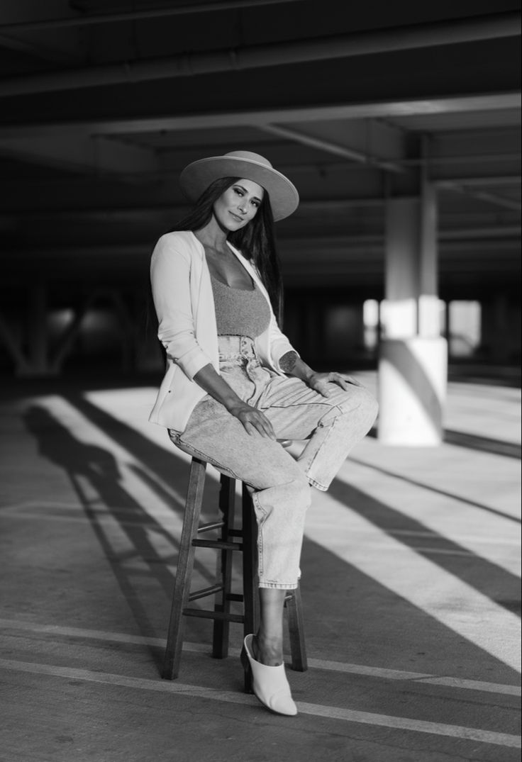 a woman with long dark hair is sitting on a wooden chair in a parking garage. she is wearing a white blazer, jeans, a white hat, and white shoes. the photo is in black and white. the sun is shining on her, creating shadows and highlighting the stripes on the floor. the background is a parking garage, and you can see the concrete columns and the ceiling. the woman is looking at the camera.