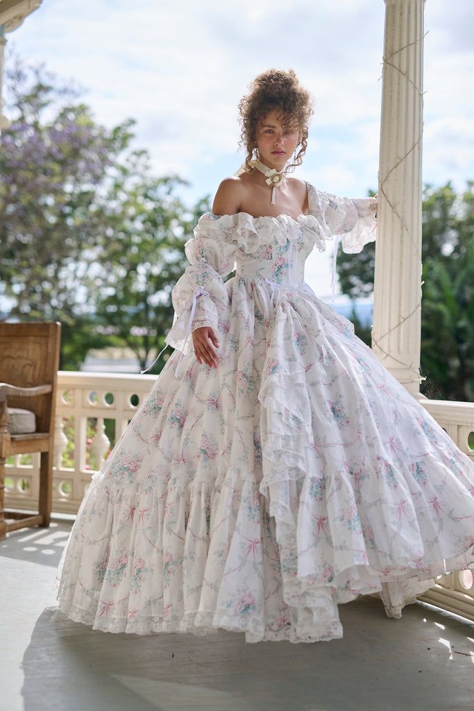 a woman in a white floral gown stands on a porch. the image is taken from eye level and the lighting is natural. the overall feel is romantic and vintage.