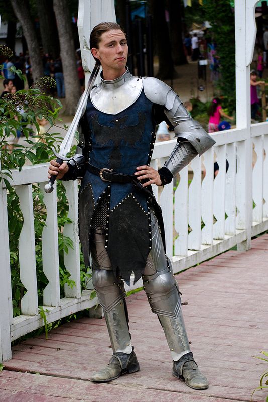 young man dressed in a medieval knight costume stands on a bridge, holding a sword. he is wearing a suit of silver armor, with a blue tunic underneath. his armor includes chainmail, a helmet, and knee and shin guards. the sword is long and slender, held casually over his shoulder.  he is standing with one hand on his hip and looks directly at the viewer with a somewhat serious expression. the bridge is white and wooden, and the background shows trees, greenery and other people (blurred), suggesting the setting is a renaissance fair or similar historical event.