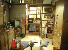 this is an image of a cluttered living space with traditional japanese style. there is a small kitchen visible at the back of the room. the predominant color palette consists of browns, beiges, and tans. the lighting suggests it is daytime and the light is coming from the window at the back of the room.