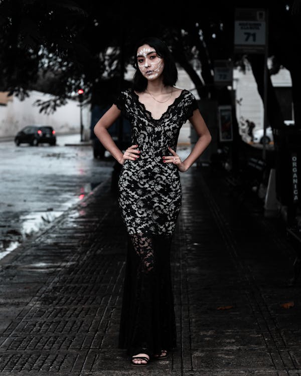 a full shot of a woman standing on a wet city street at night. she's wearing a long black lace dress and has intricate skull makeup on her face, suggestive of dia de los muertos (day of the dead) style. the atmosphere is dark and moody, and she appears confident and elegant in her pose. the background is blurred, highlighting her as the focal point.  the overall aesthetic is stylish and slightly gothic.