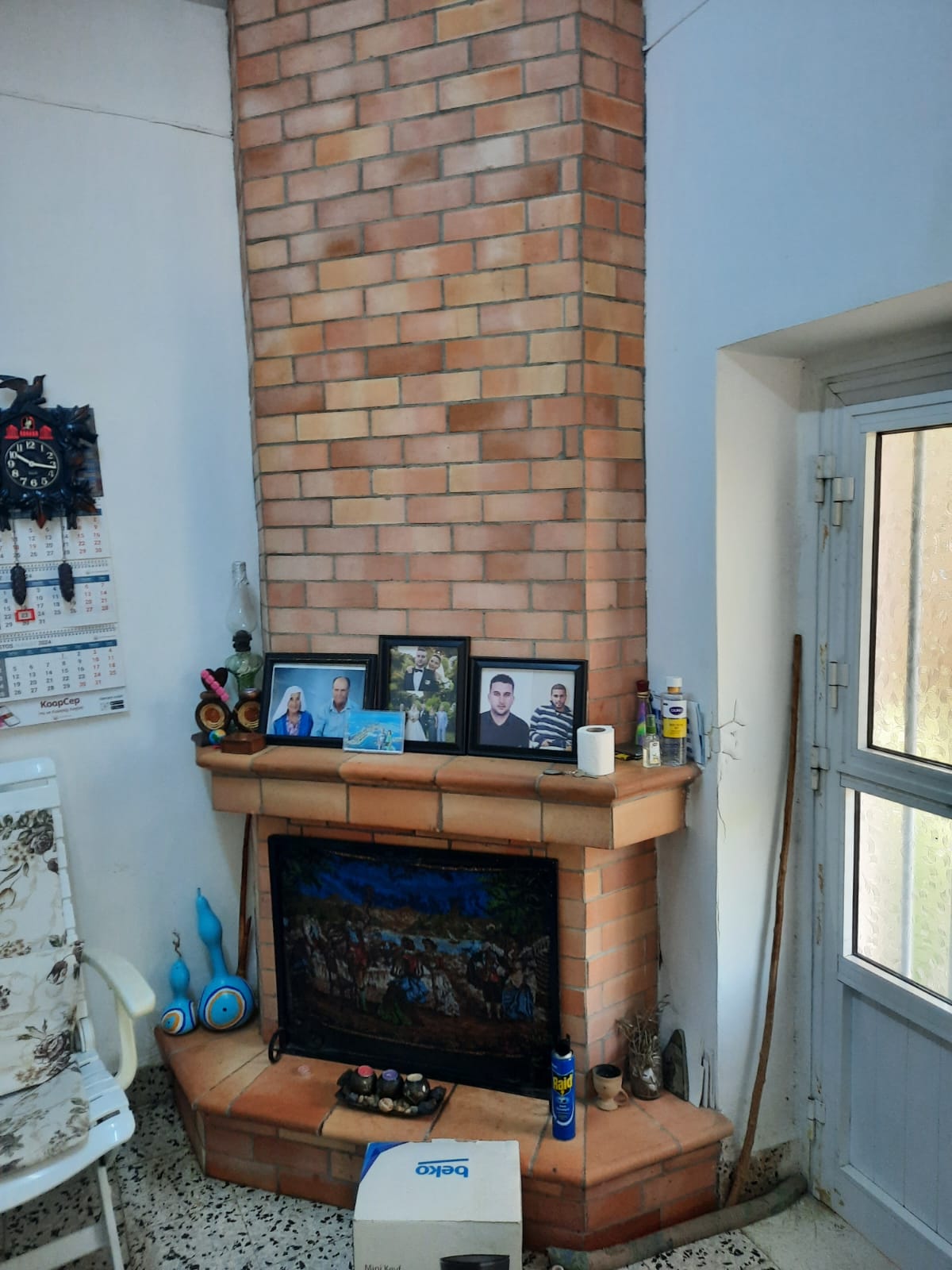 the image shows a brick fireplace with a mantle, adorned with photos, decor items, and a roll of toilet paper. a chair sits to the left of the fireplace. a door is to the right of the fireplace. a clock and calendar are hanging on the wall near the chair. there's a box in front of the fireplace.