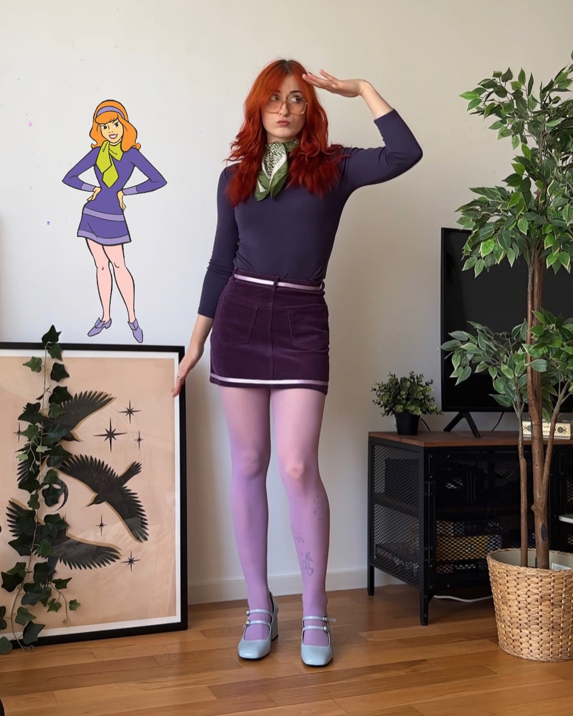 a fulllength photograph depicts a woman cosplaying as daphne blake from scooby doo. she's wearing a purple longsleeved top, a purple corduroy miniskirt, lilac tights, and light blue mary jane shoes.  she also sports a green scarf and glasses. her long red hair is styled loosely. in the background is a framed illustration of daphne from the cartoon, along with indoor plants and a darkgrey metal media cabinet. the overall aesthetic is retroinspired and reminiscent of the 1970s.