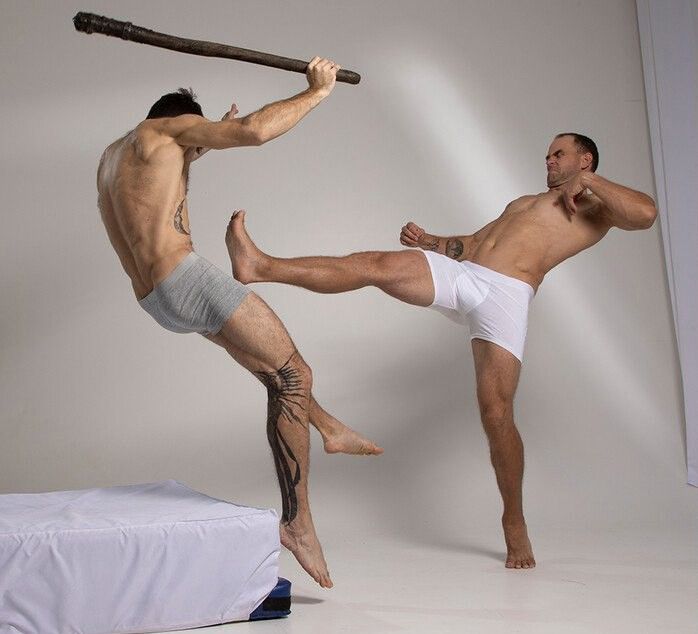 two men in white underwear are in a martial arts fight. one man is kicking the other man who is holding a stick. the men are bare chested and  the man in the foreground has a tattoo on his leg. the background is white.