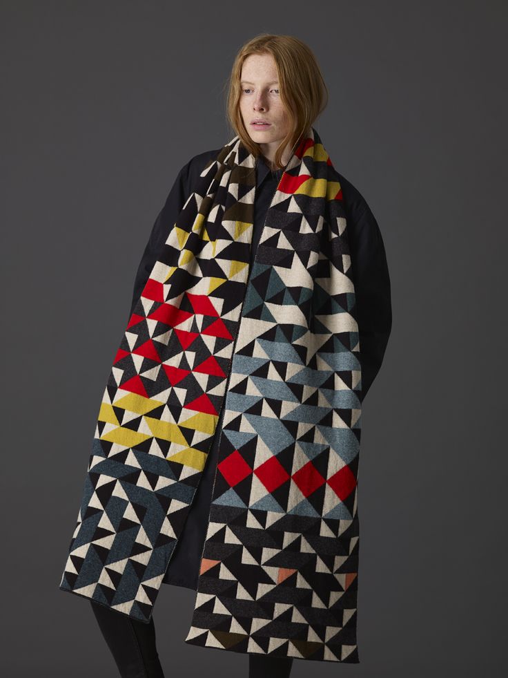 a young woman with long straight red hair is wearing a black coat and a long, colorful patterned scarf. the scarf is a geometric design with black, white, red, yellow, blue and green. the woman is standing in a studio on a gray backdrop.