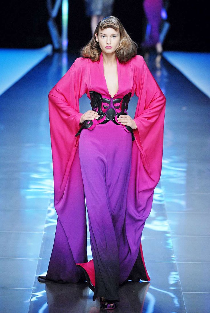 a model walks down a runway in a pink and purple ombre long dress with long sleeves and a black bodice. she has short brown hair and dramatic makeup.