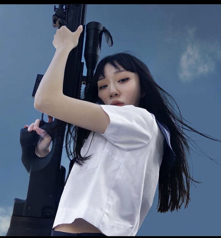 a woman with long black hair is holding a large black gun with a scope. she is wearing a white shortsleeved shirt that appears to be part of a school uniform. the background is a blue sky with some clouds. the woman is looking directly at the camera.