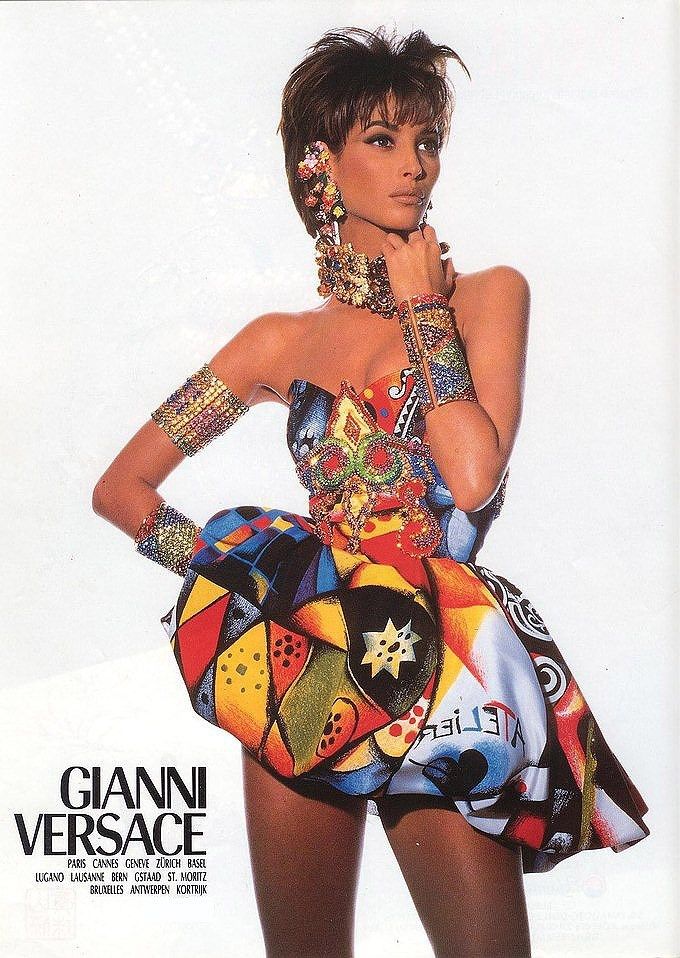 a vintage gianni versace advertisement featuring model linda evangelista. she is wearing a strapless, short dress with a bold, colorful pattern. evangelista is adorned with statement jewelry including large earrings, a choker necklace, and ornate bracelets. the ad also includes the gianni versace logo and a list of city locations.
