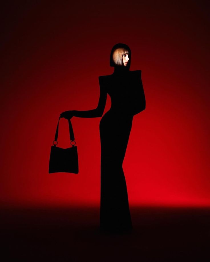 a woman in a black dress and handbag stands in front of a red background. the light is positioned behind her, creating a silhouette effect.