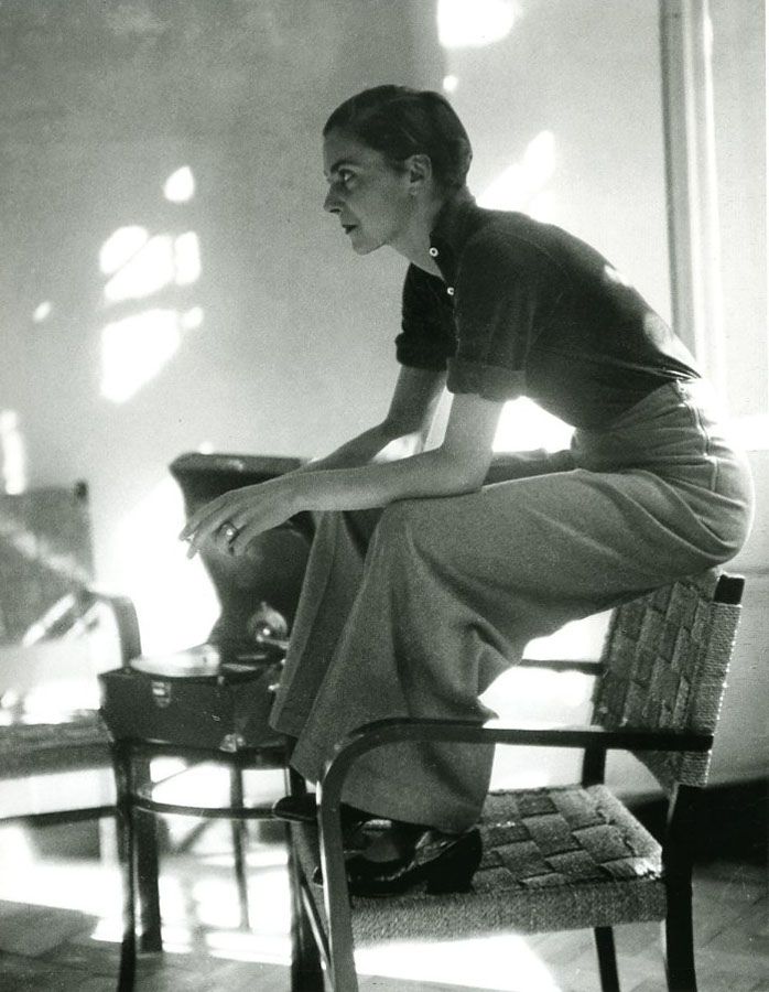 a black and white photograph of a woman sitting on a chair. she is wearing a dark shirt, pants, and shoes. she is looking to the left of the frame. the photograph is likely from the 1940s.
